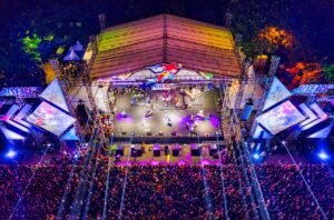 A spectacular aerial view of a lively nighttime outdoor concert with a large crowd and colorful stage lighting.